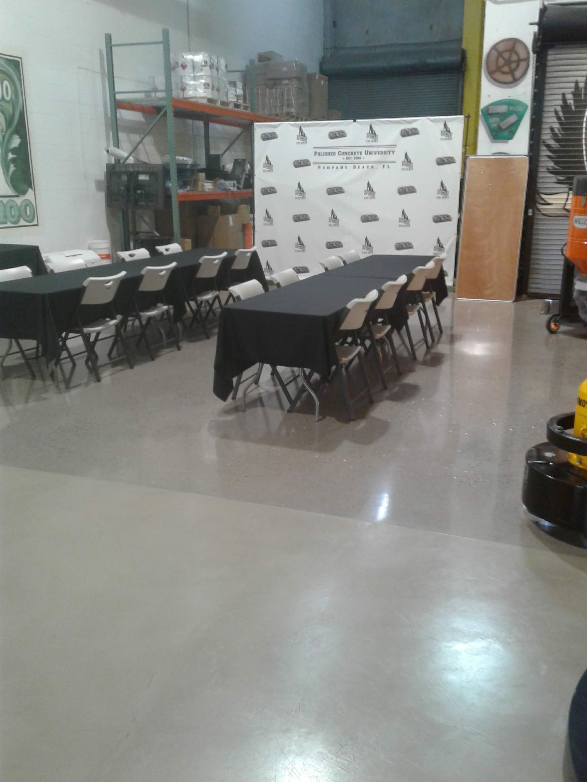 A workshop or event space featuring two rows of long tables with black tablecloths and chairs, facing a branded backdrop.