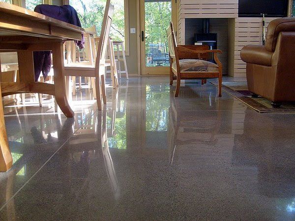 A sunlit room featuring polished grey concrete floors that reflect the wooden dining furniture and leather armchair.