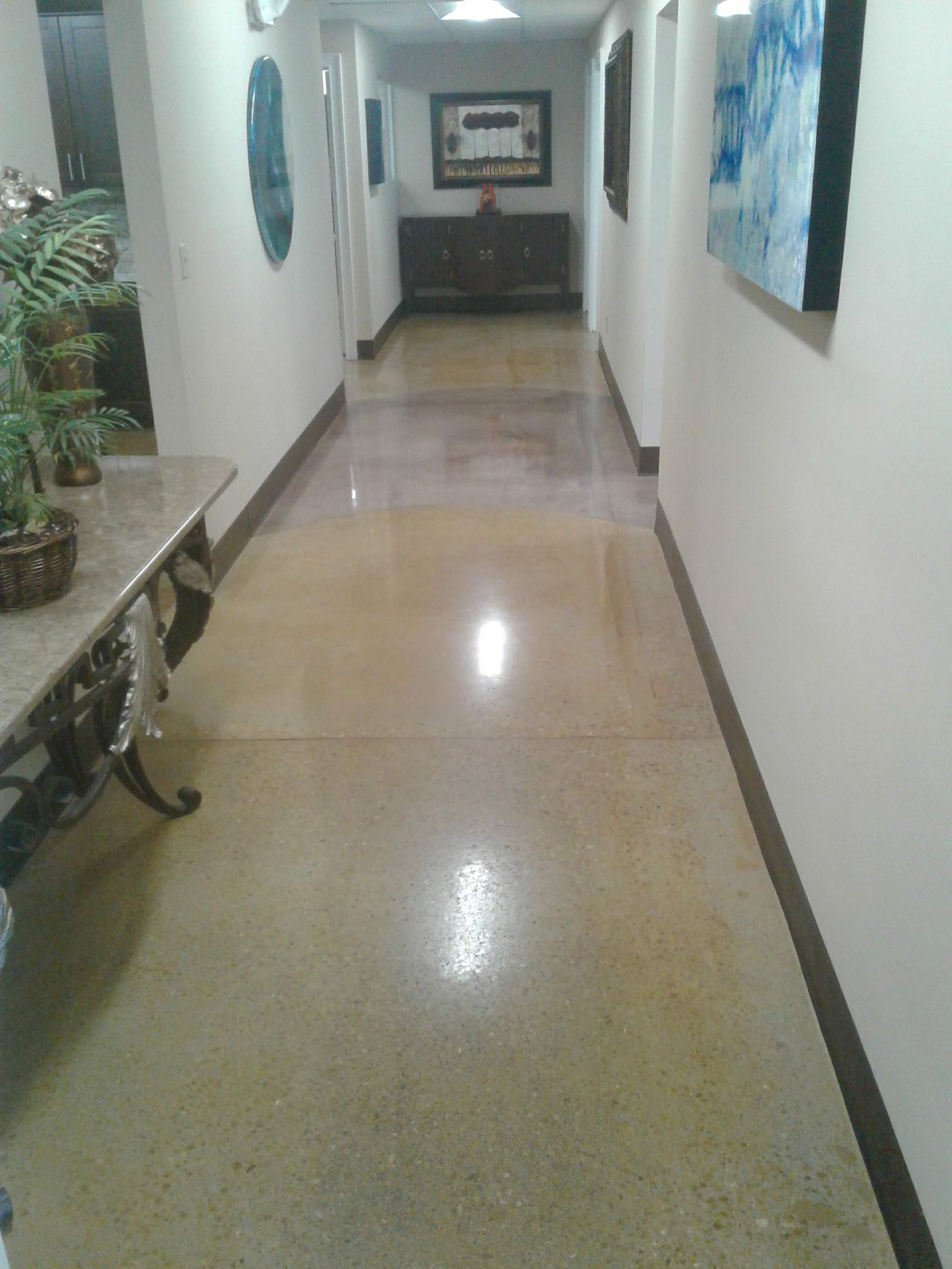 A brightly lit residential hallway with polished stone floors, decorative artwork, and a console table.