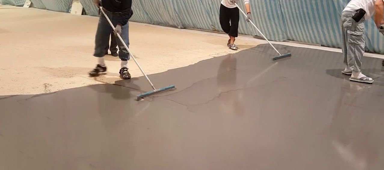 Workers use long-handled squeegees to spread wet, gray concrete across a floor in a large, open space.