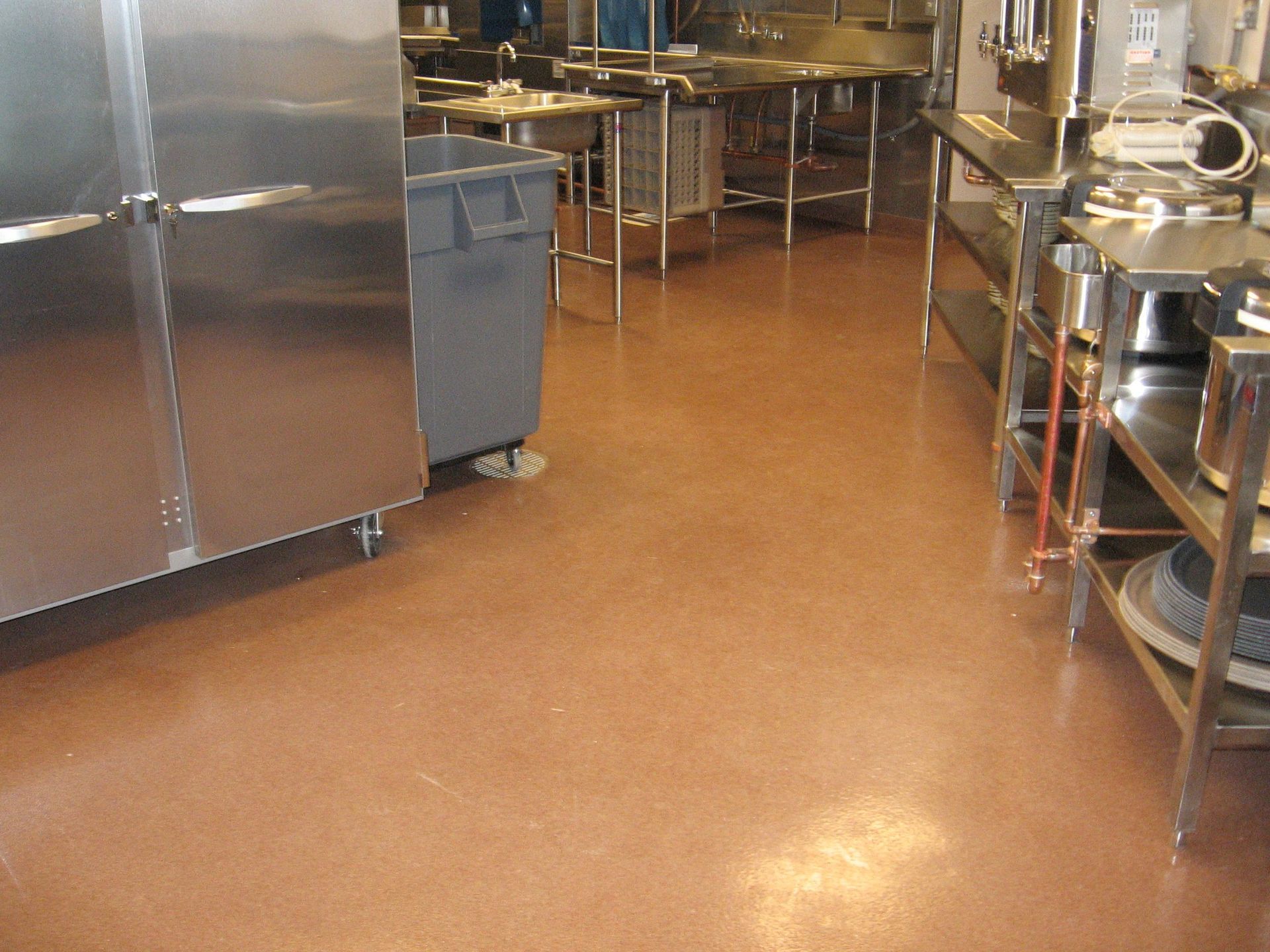 An industrial kitchen with stainless steel equipment, a large gray trash bin, and a light brown floor.