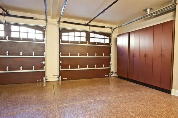 An organized garage with two closed dark wood carriage-style doors, speckled flooring, and built-in matching cabinets.