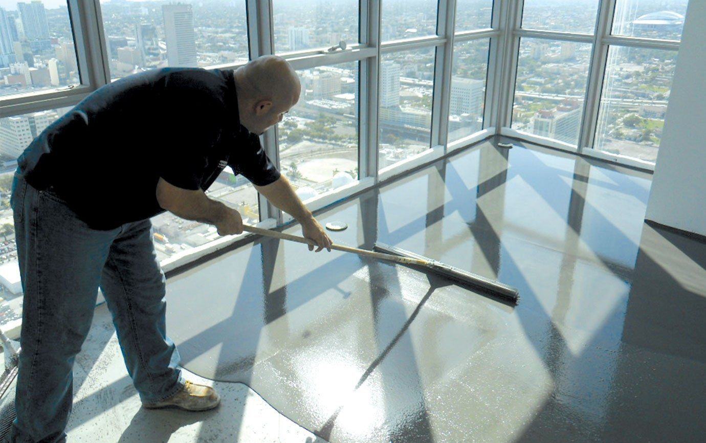 A person uses a squeegee to spread a wet, leveling compound across a concrete floor in a room with floor-to-ceiling windows.