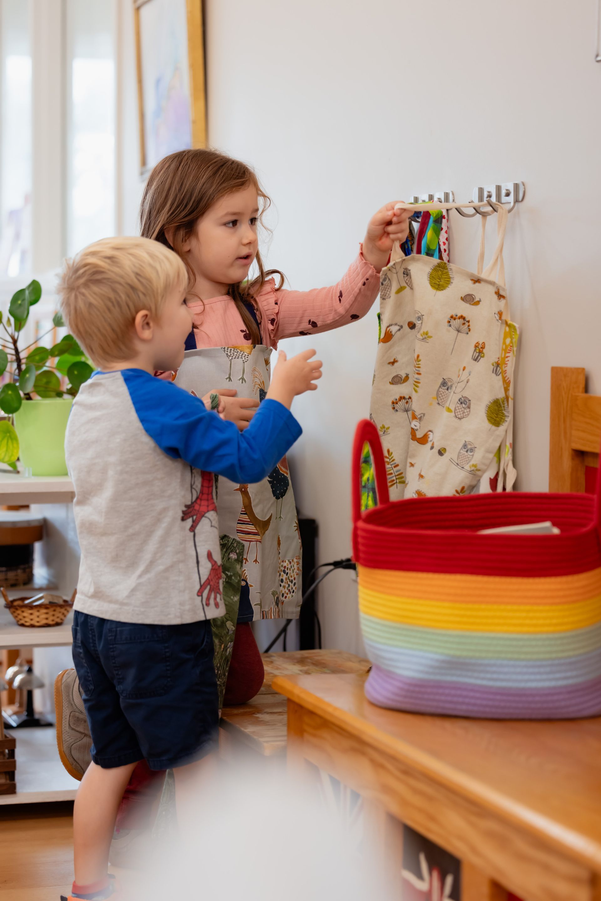 Two children hanging colorful clips on a rack beside a striped basket in a bright room