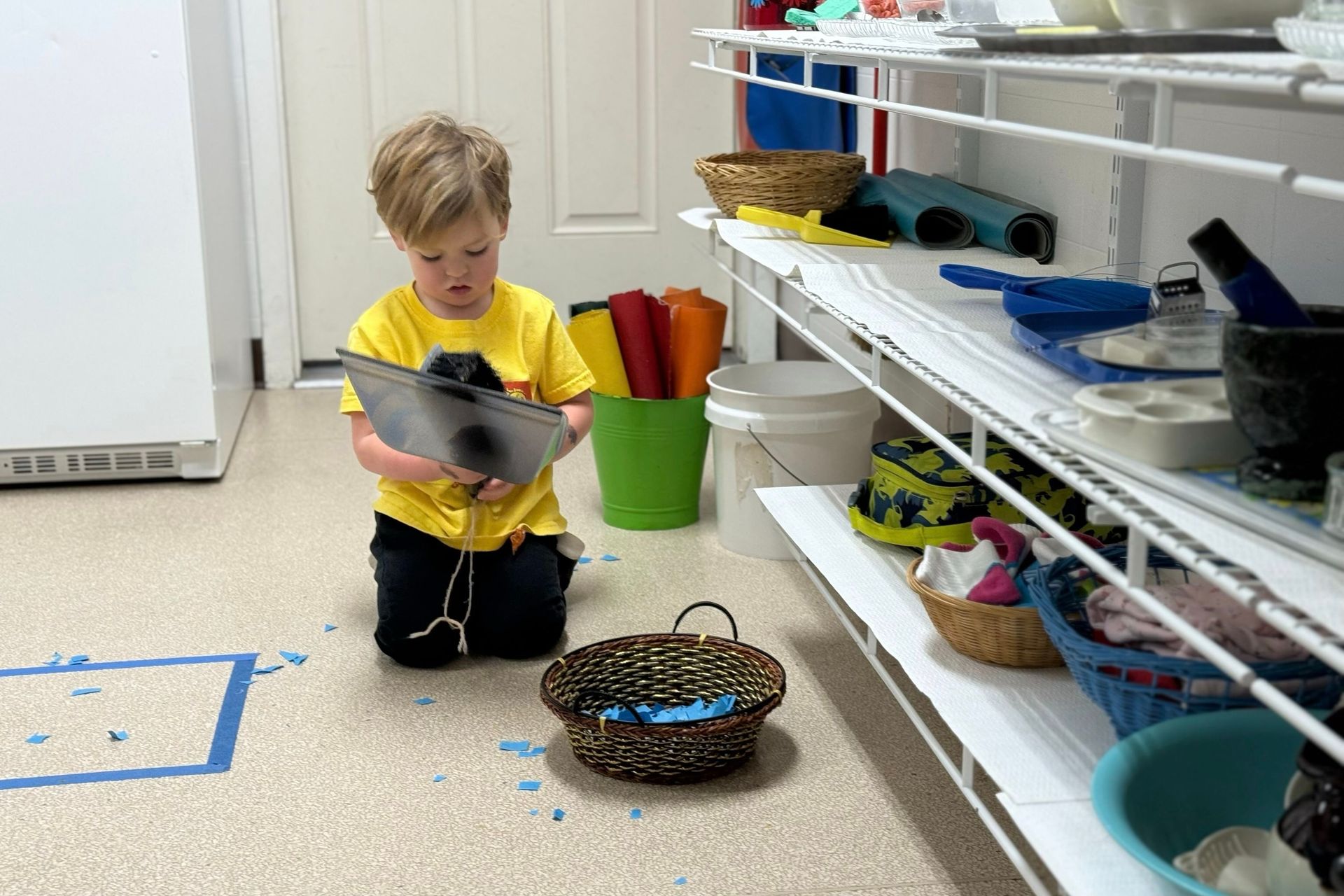 Child kneeling on the floor with a tablet in a classroom, next to shelves of colorful baskets and supplies