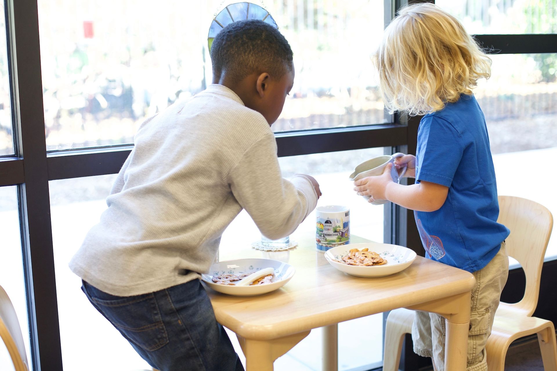 Two children sharing a snack at a small table by a bright window