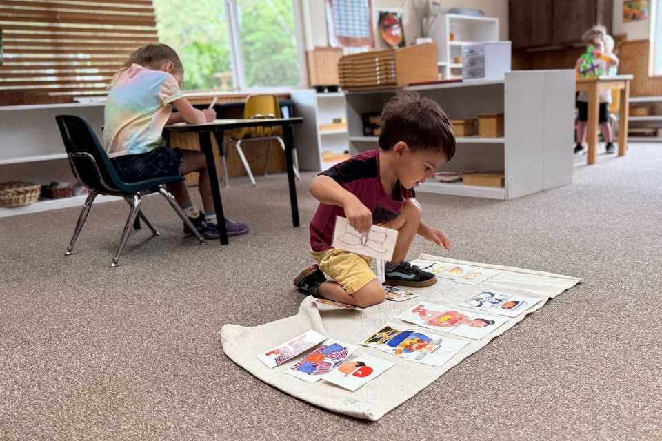 Boy playing with cards on floor in a  Montessori classroom, another child at a desk.