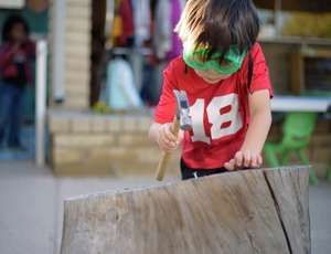Child in red shirt hammering wood stump outside.