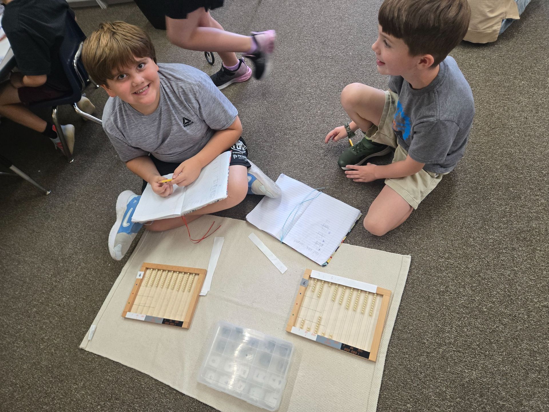 Two students work on a school project, sitting on a rug with wooden counting frames, notebooks, and supplies.