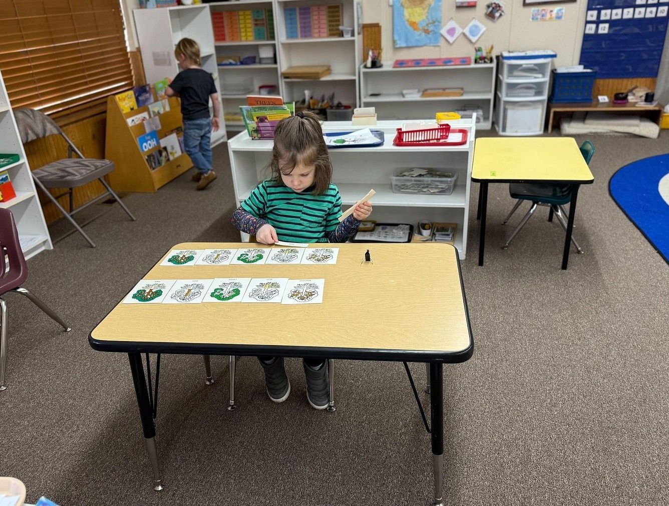 A student in a green striped shirt works with learning cards on a classroom table while another student stands nearby.