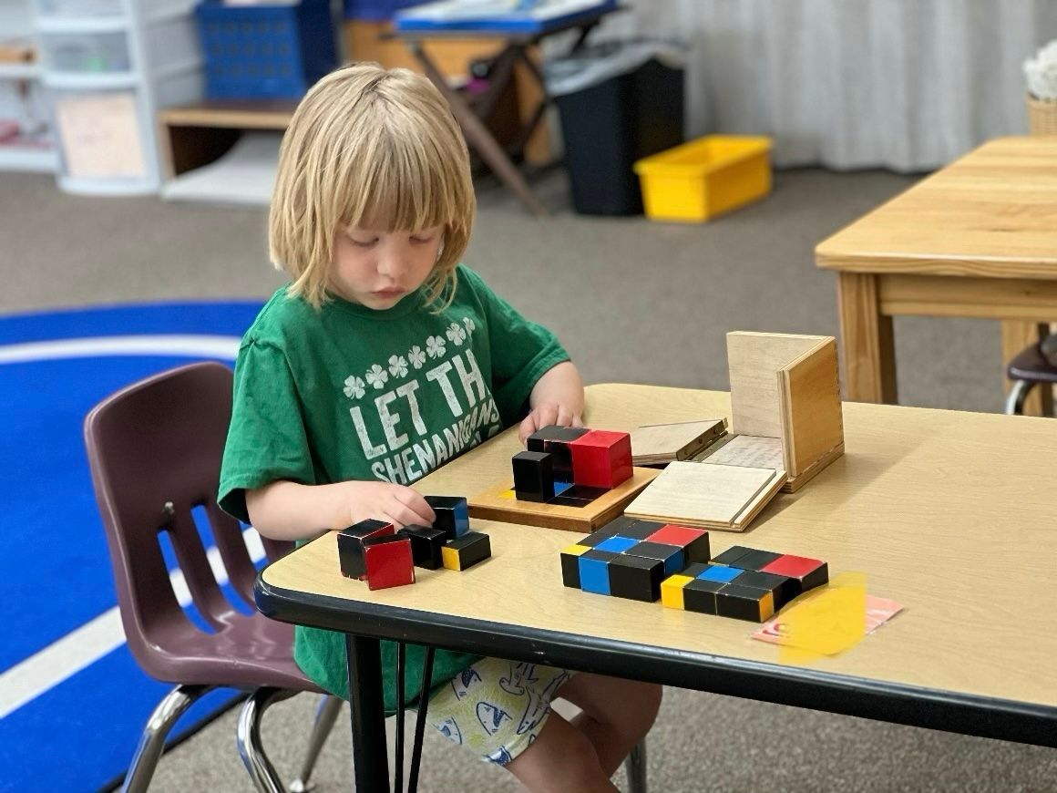 Young child working with Montessori trinomial cube at a table in a classroom.