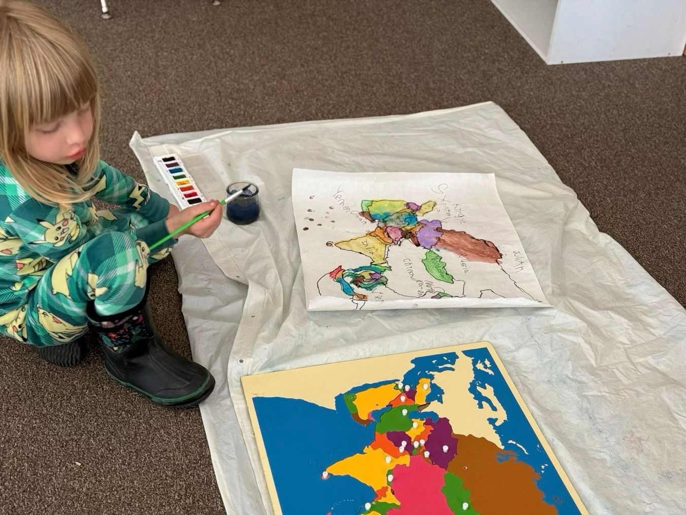 Child painting on a white sheet beside colorful abstract artwork on the floor