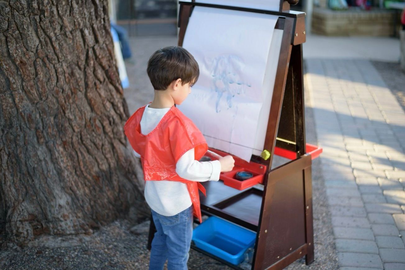 Child painting at an easel outdoors beside a tree and paved walkway