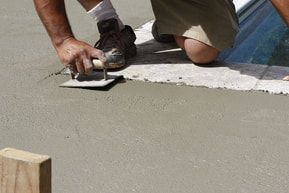 A man is kneeling down and using a trowel to spread concrete on a sidewalk.