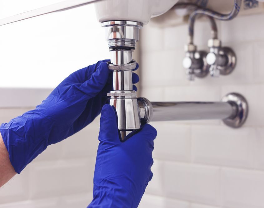 A plumber wearing blue gloves unscrewing pipes below a sink.