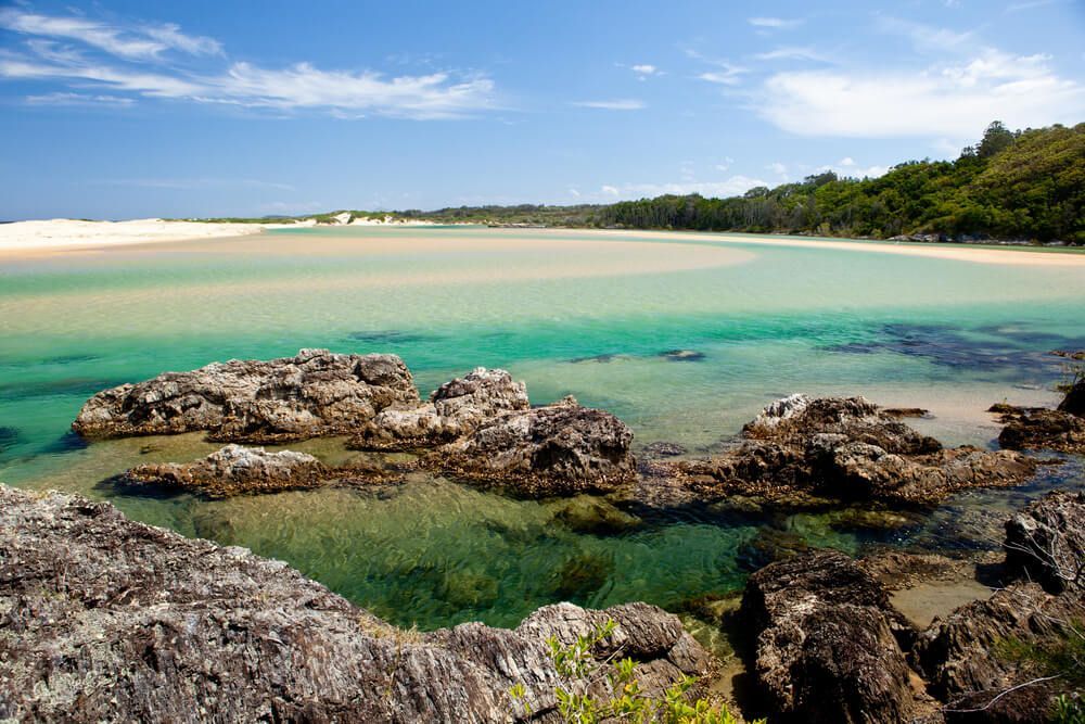 A Large Body Of Water With Rocks In The Foreground And A Beach In The Background — Coastal Plumbing Group in Sawtell, NSW