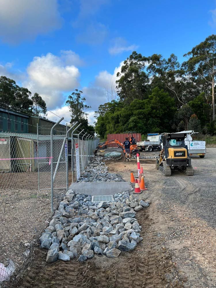 A Bulldozer Is Driving Down A Dirt Road Next To A Pile Of Rocks — Coastal Plumbing Group in Woolgoolga, NSW