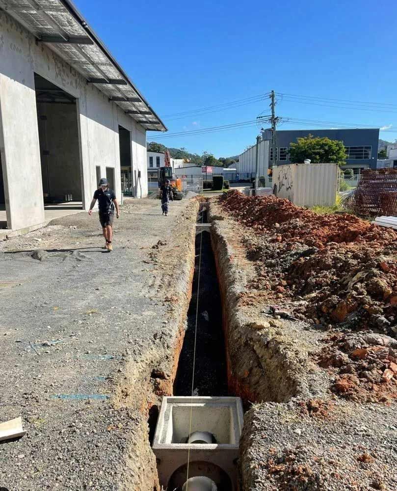 A Man Is Walking Down A Dirt Road Next To A Building Under Construction — Coastal Plumbing Group in Woolgoolga, NSW