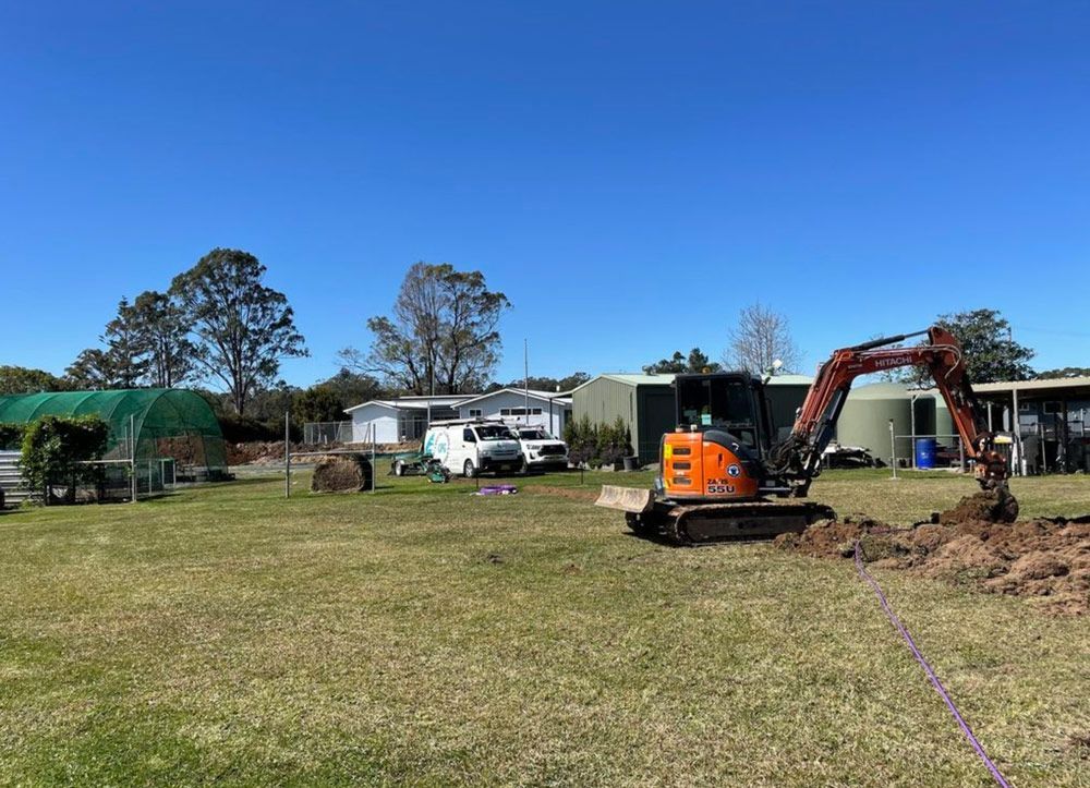 A Small Excavator Is Digging A Hole In A Grassy Field — Coastal Plumbing Group in Woolgoolga, NSW