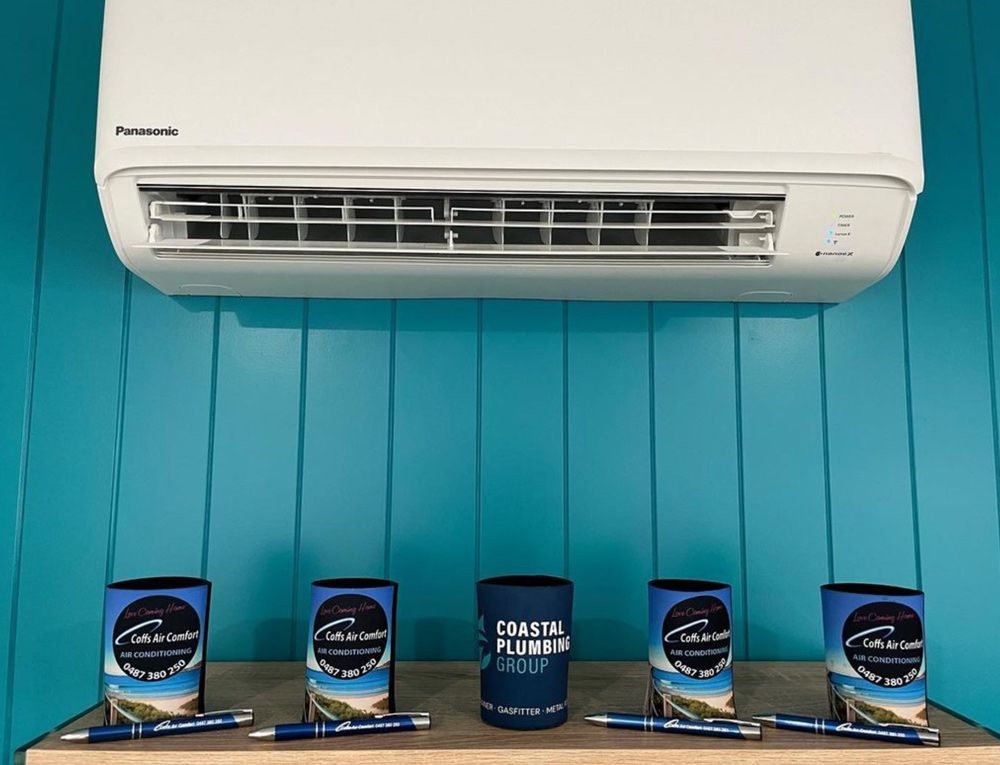 A White Air Conditioner Is Hanging On A Blue Wall Next To A Table With A Row Of Coolers And Pens — Coastal Plumbing Group in Woolgoolga, NSW