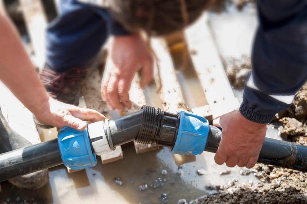 Two Workers Connect Plastic Pipes With Blue Fittings in a Muddy Area
