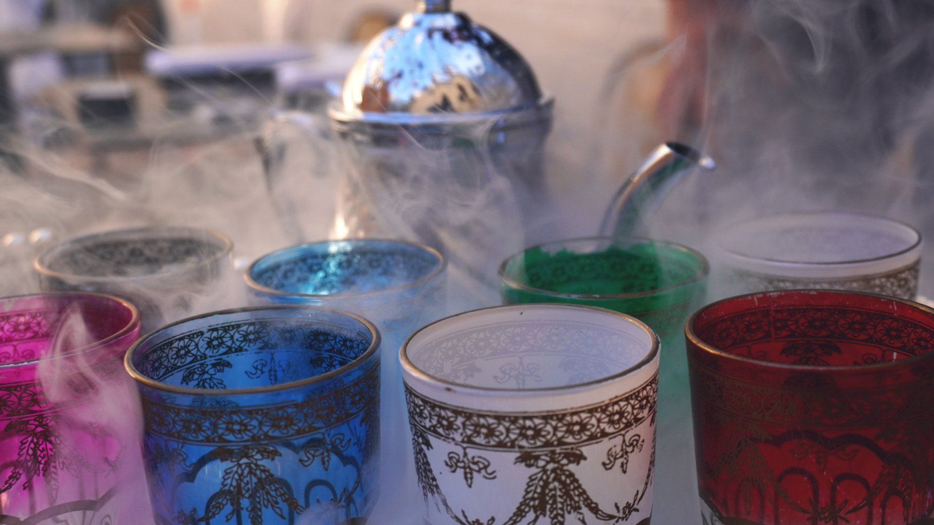 Un gruppo di bicchieri colorati sono appoggiati su un tavolo e da essi esce del fumo.