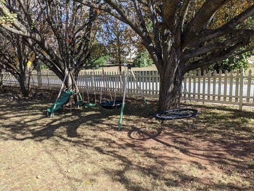 Backyard with large trees, playground swings, and a white picket fence in sunlight