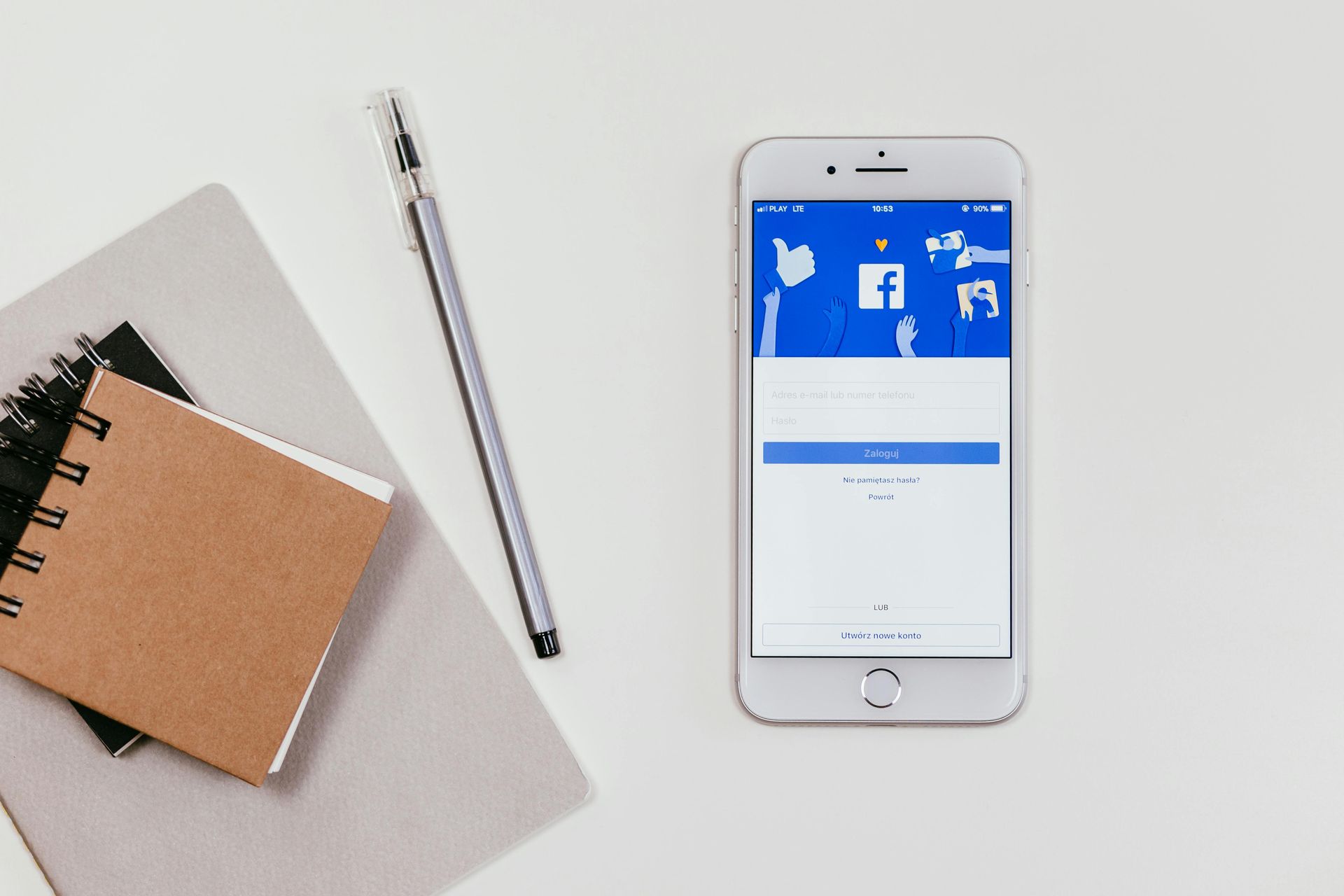 Smartphone displaying a Facebook login screen beside a notebook, pen, and laptop on a desk