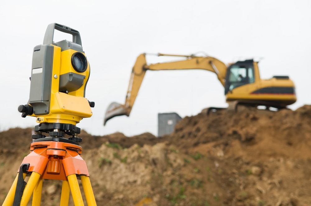 A Construction Site With A Yellow Excavator In The Background And A Theodolite In The Foreground  — CalCo Surveyors Pty Ltd in Gloucester, NSW