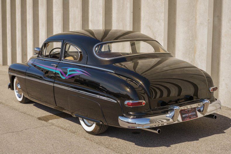 A black, vintage custom Mercury coupe with whitewall tires and colorful side graphics, parked against a ribbed wall.