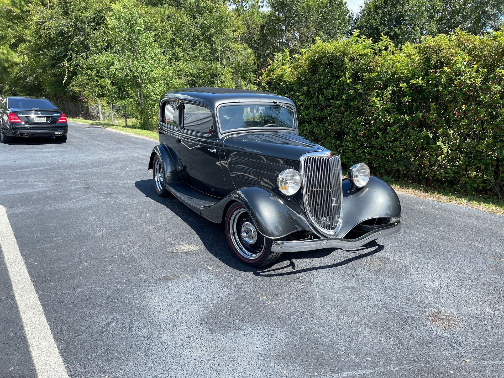 A vintage, dark gray, four-door sedan parked on an asphalt lot in front of a green hedge, with a modern car behind it.