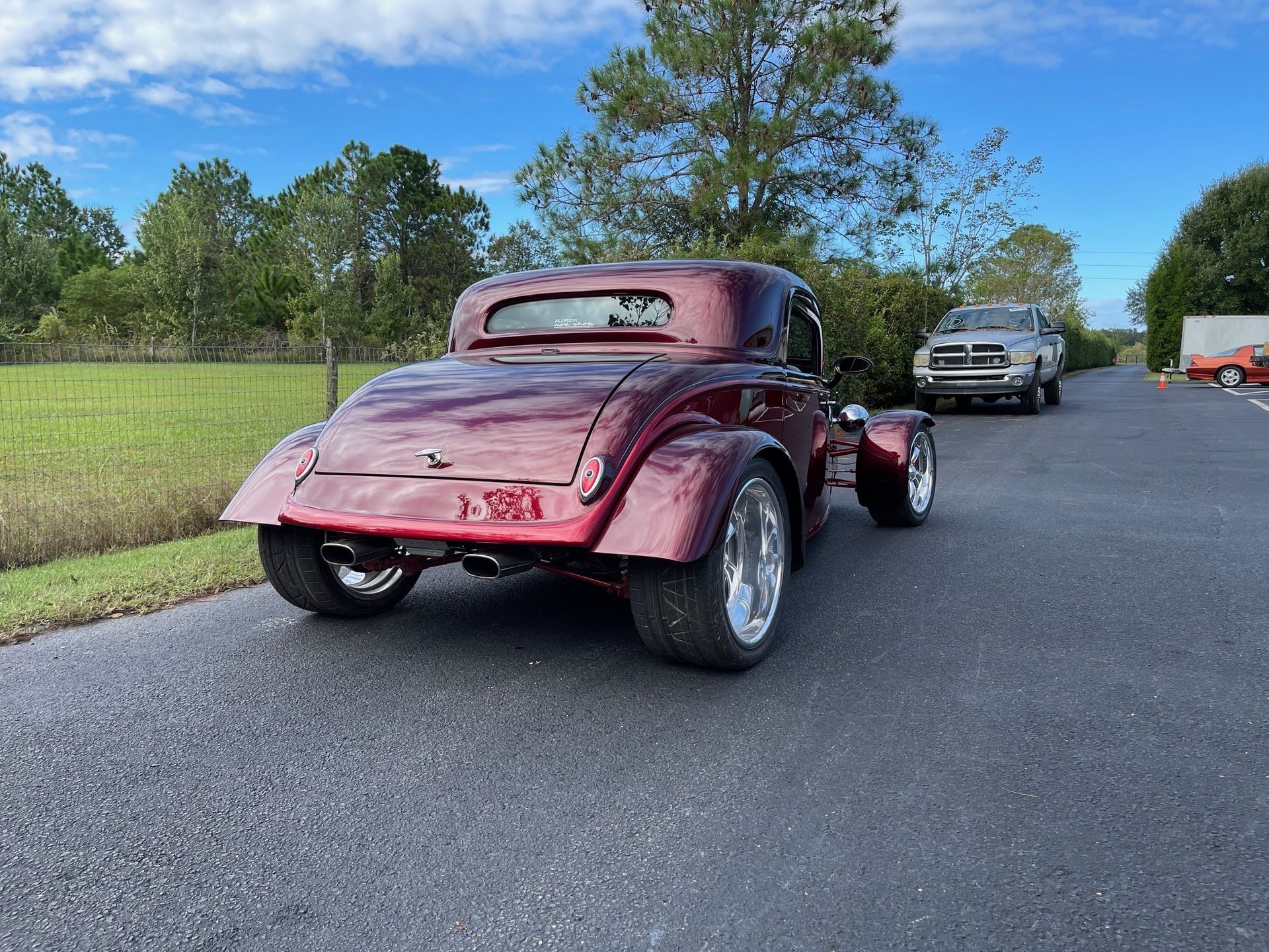 A dark cherry-red custom hot rod coupe parked on an asphalt road with trees in the background.