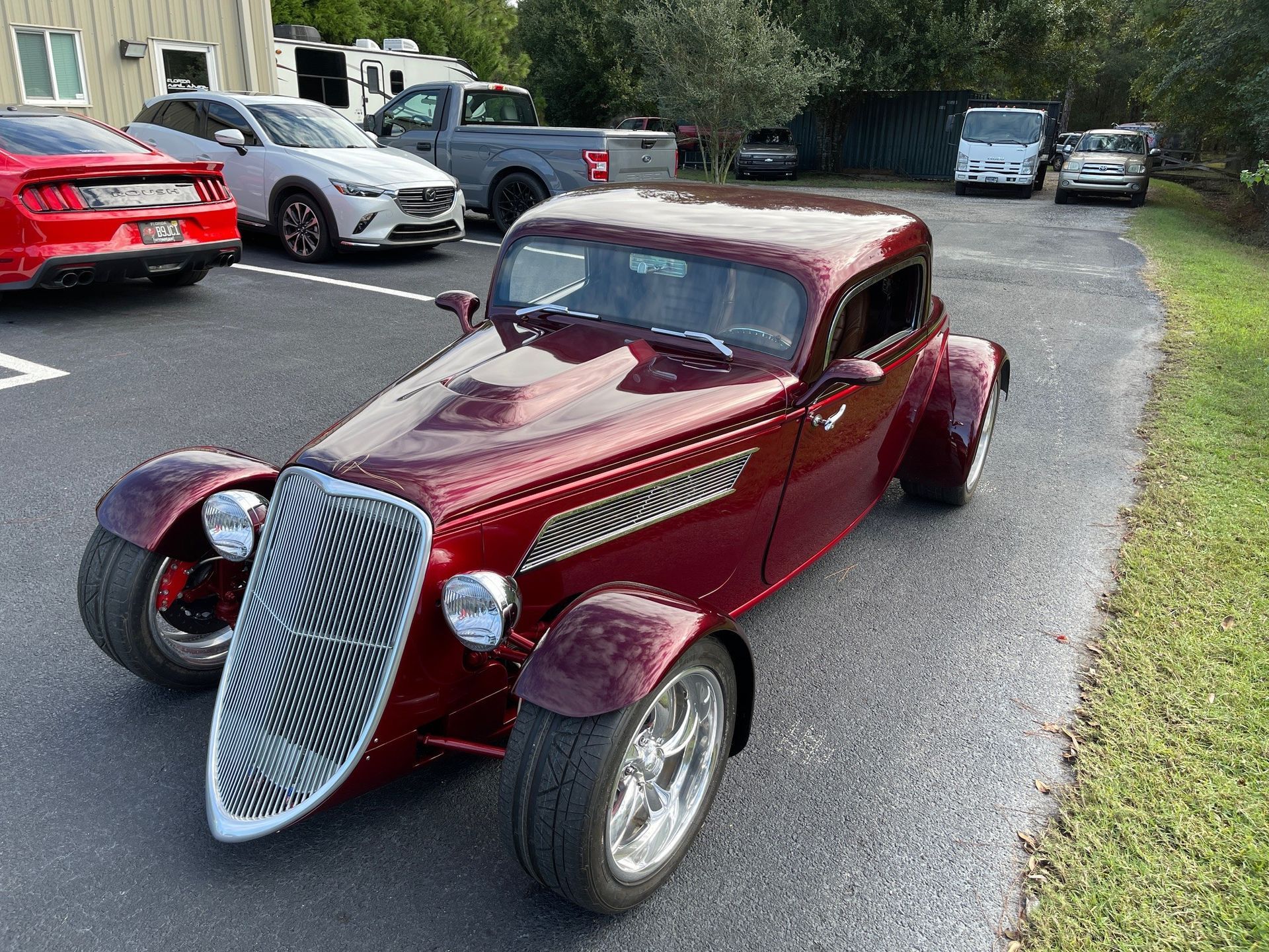 A custom burgundy hot rod with a tall chrome grille parked in an asphalt lot with other vehicles in the background.