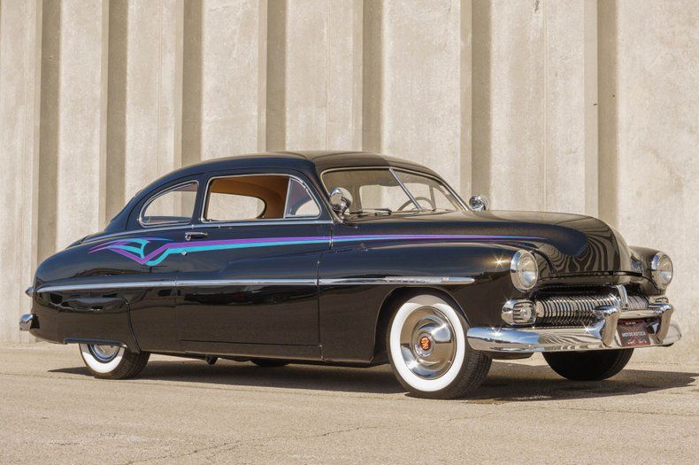 A classic black custom Mercury coupe with wide whitewall tires and teal pinstriping parked against a concrete wall.