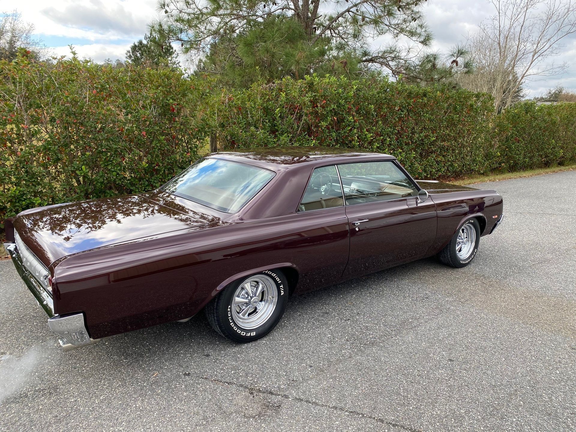 A deep maroon vintage coupe parked on a gravel driveway in front of a green hedge.
