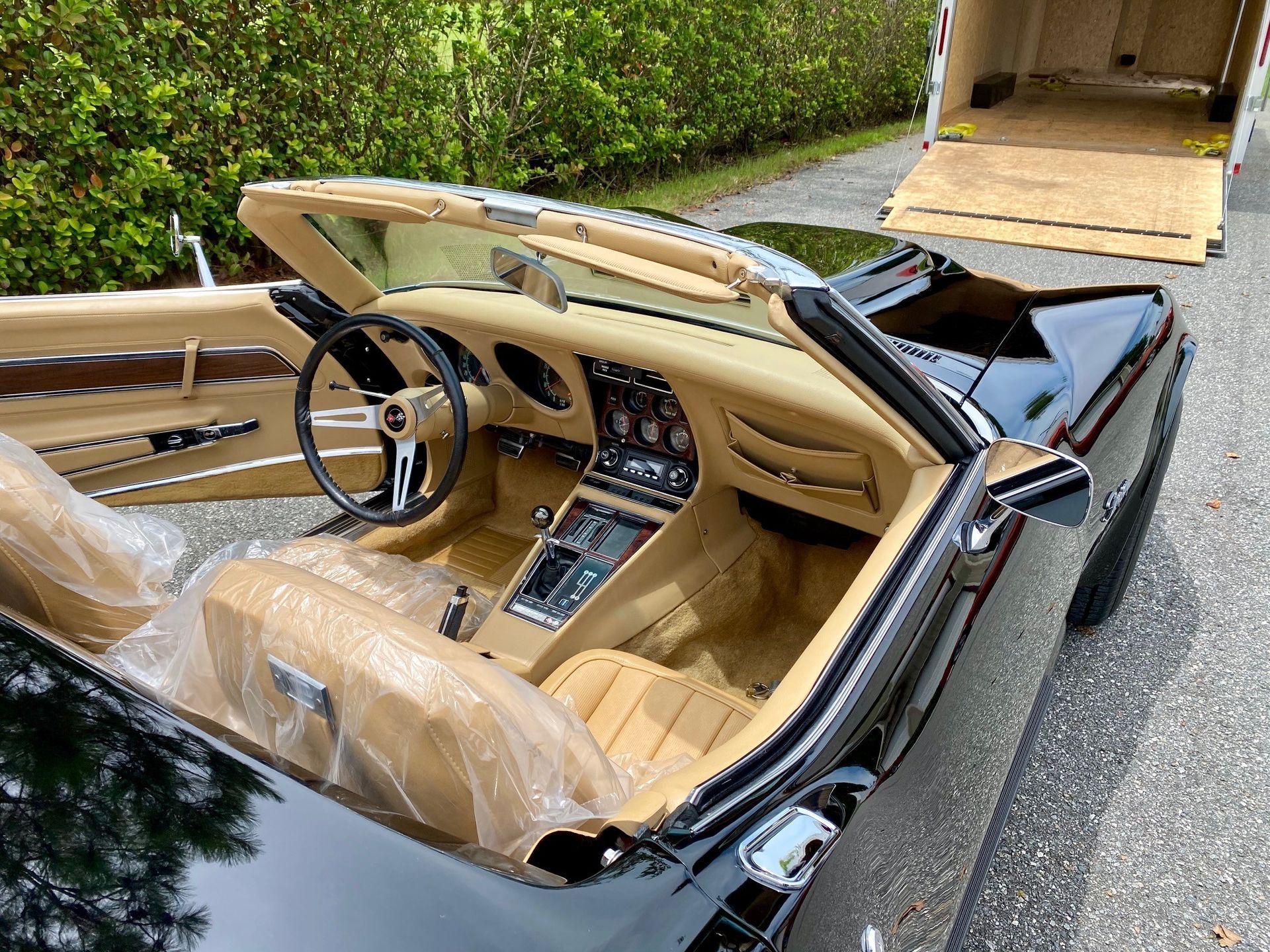A black convertible car with a tan leather interior parked near an open trailer on a gravel surface.