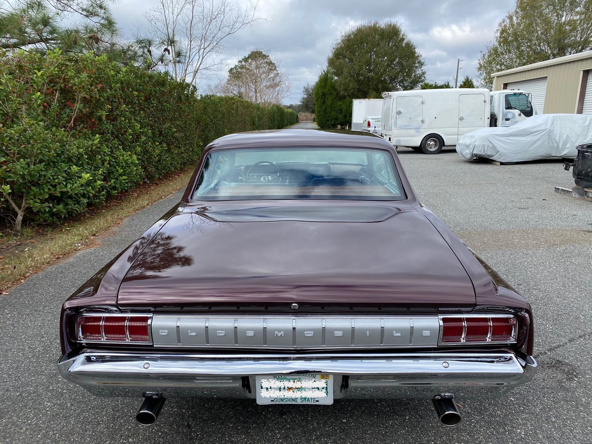 A burgundy vintage Oldsmobile car parked on a gravel lot, viewed from the rear, showing the chrome bumper and taillights.