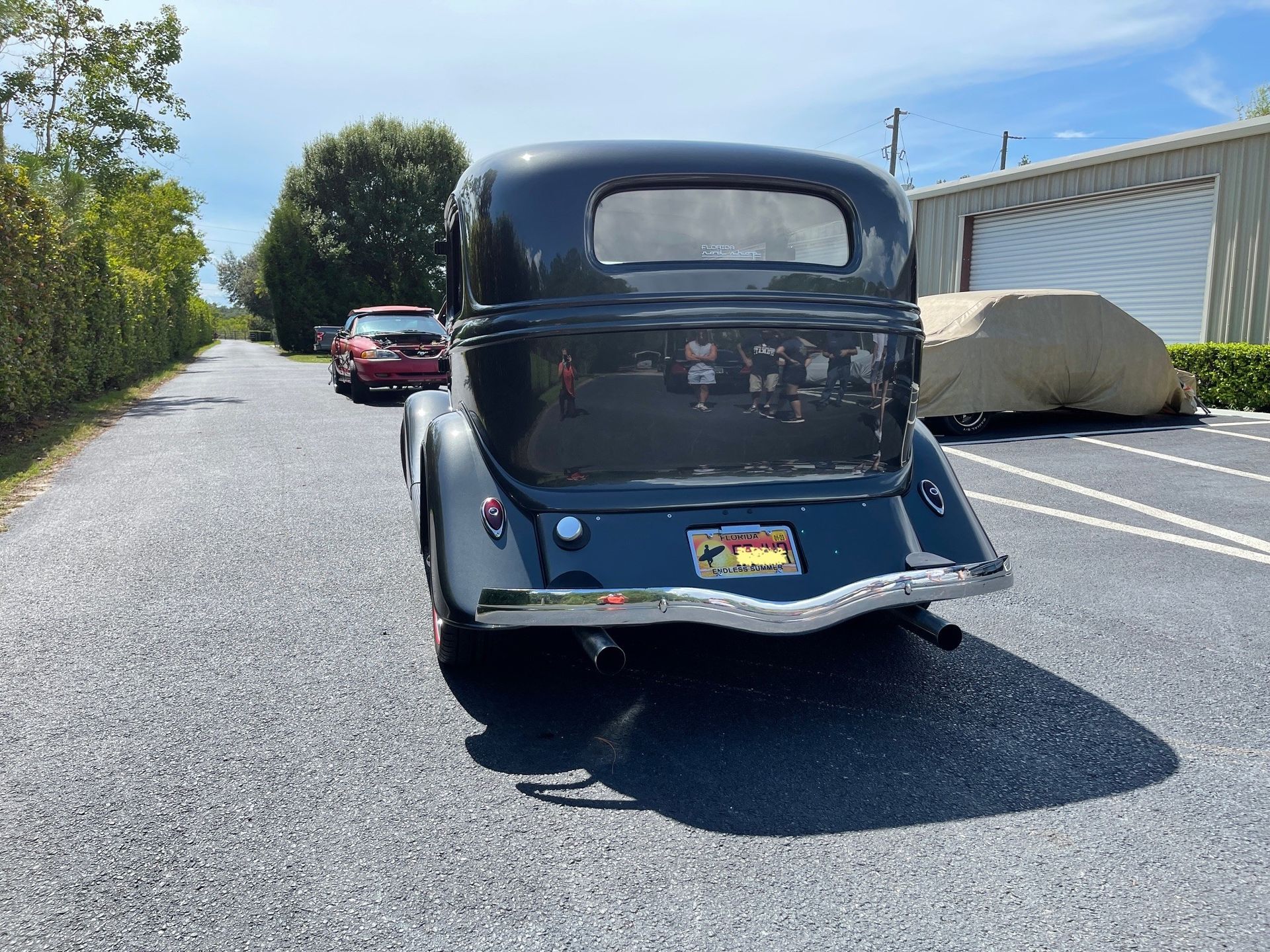 A rear view of a dark grey classic sedan parked on a gravel lot next to a covered car and another vehicle.