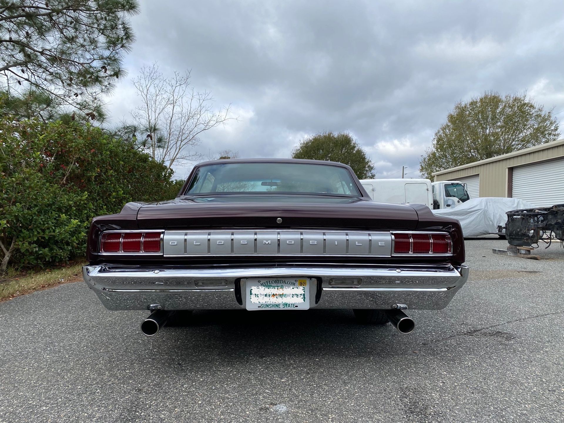 The rear of a dark-colored 1964 Plymouth, with chrome bumpers and tail lights, parked on a gravel lot under cloudy skies.