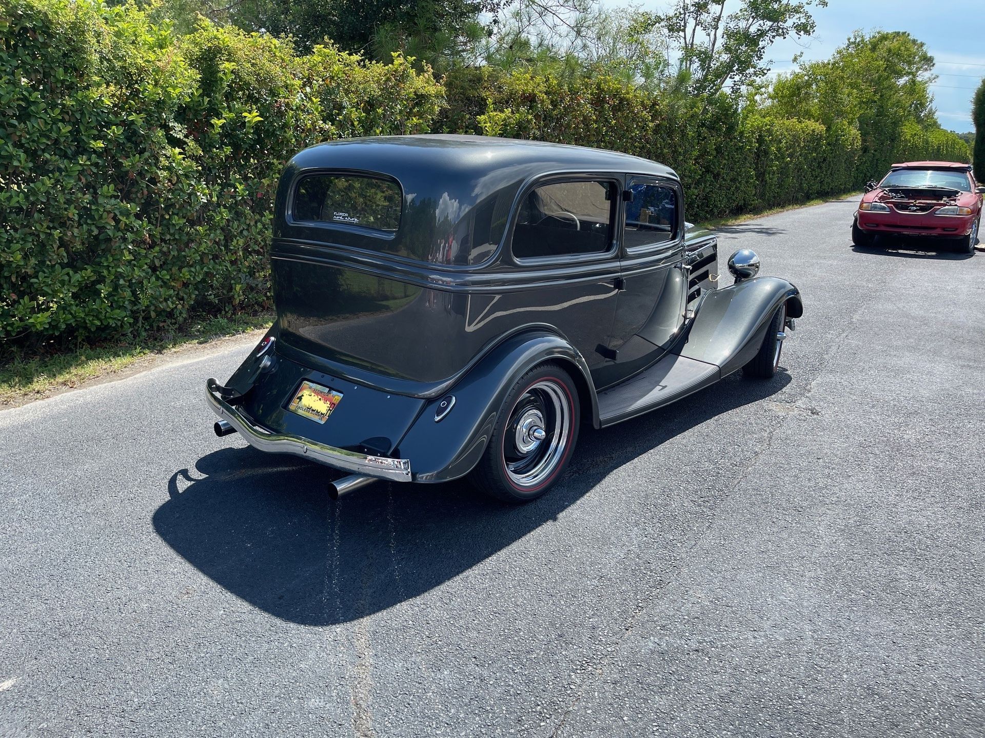 A dark grey vintage sedan parked on a gravel road, viewed from the rear, with a red car visible in the distance.