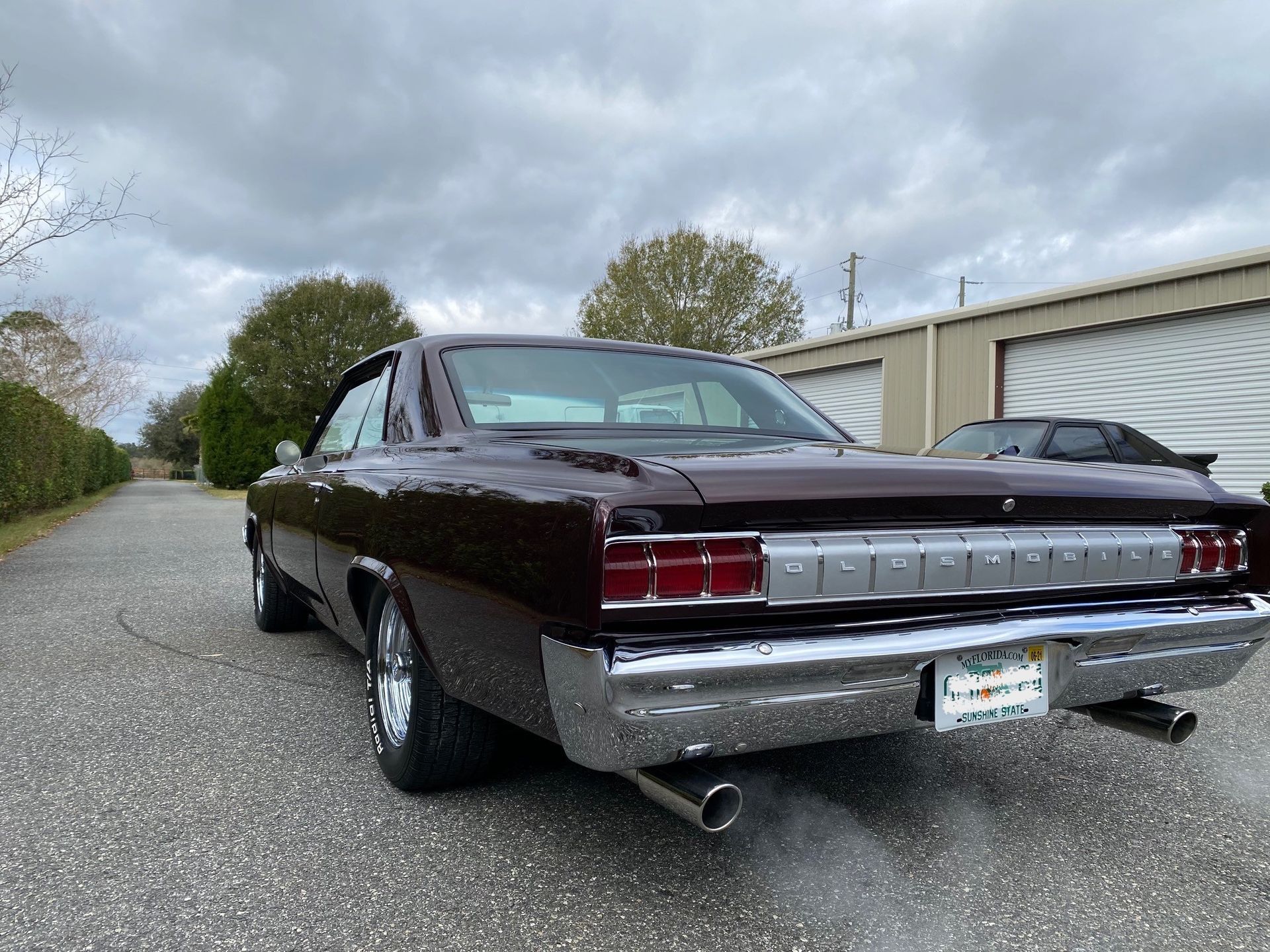 A dark maroon vintage muscle car parked on an asphalt driveway in front of storage units under a cloudy sky.