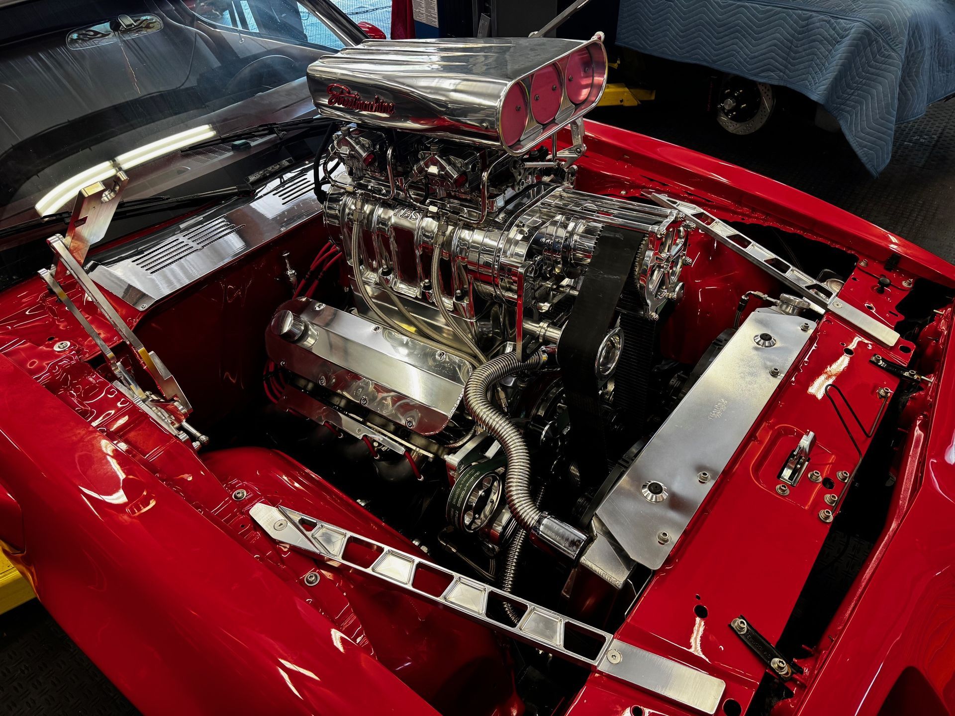 A highly detailed, chrome-plated supercharged engine sits inside the glossy, bright red engine bay of a classic car.