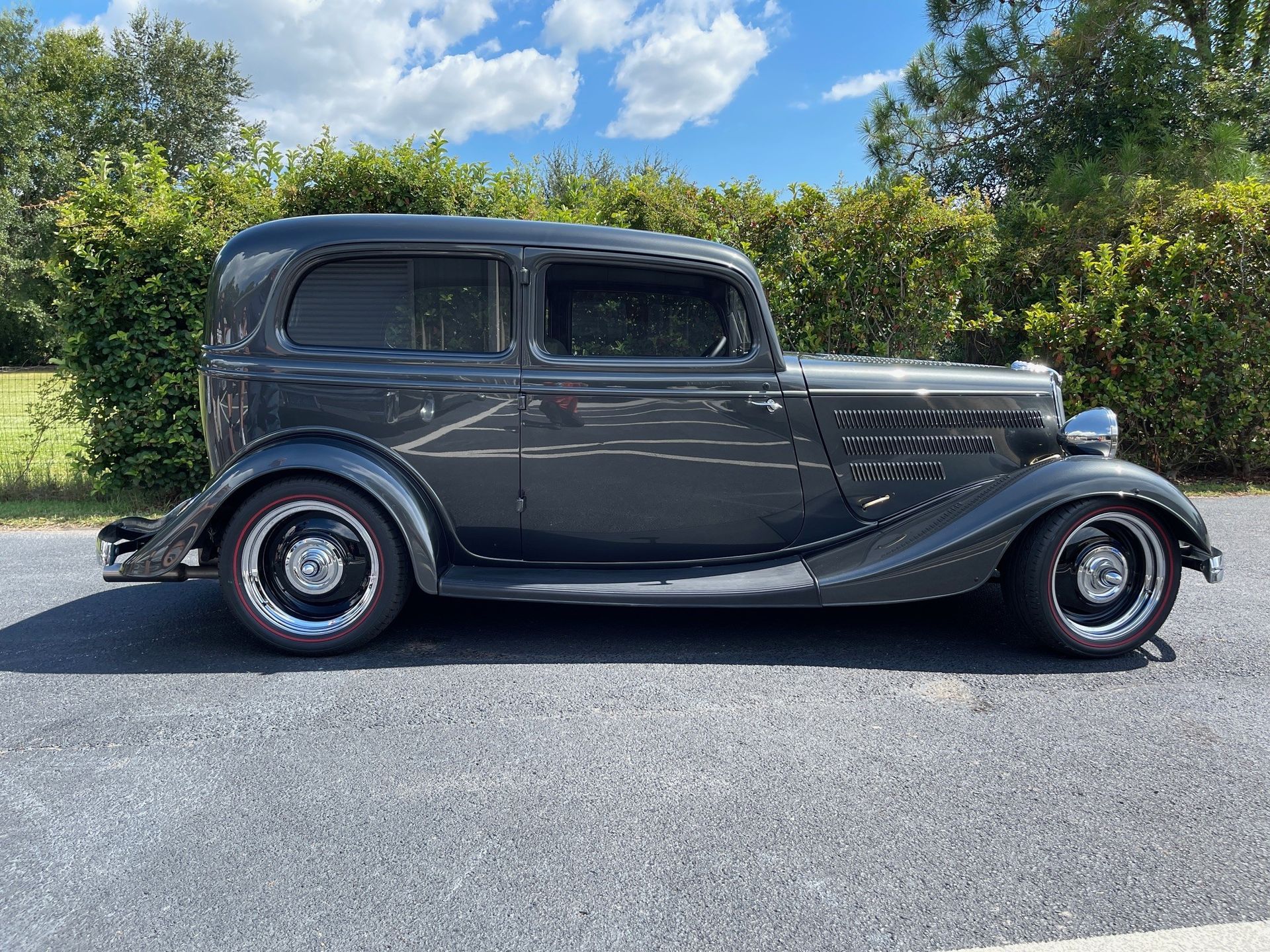 A dark gray vintage sedan parked on a gravel surface outdoors with green bushes in the background.