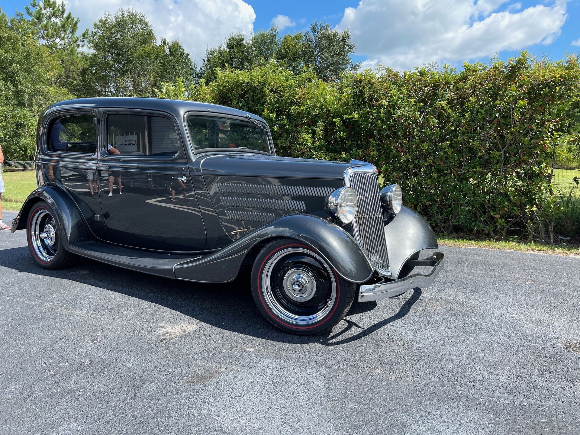 A dark gray vintage Ford sedan parked on a gravel driveway with a green hedge and blue sky in the background.