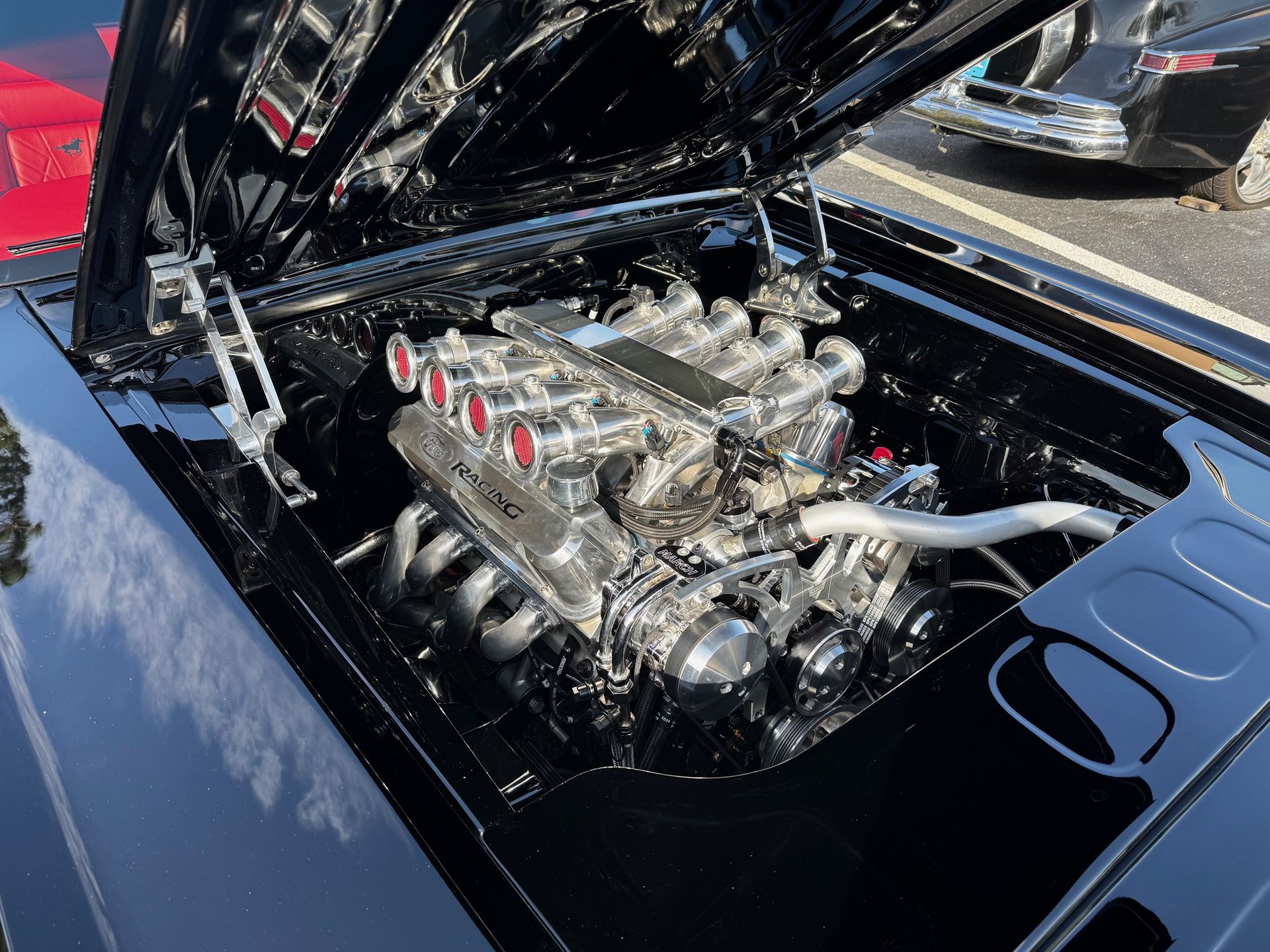 A polished black car engine bay featuring a high-performance chrome engine with red-accented velocity stacks.