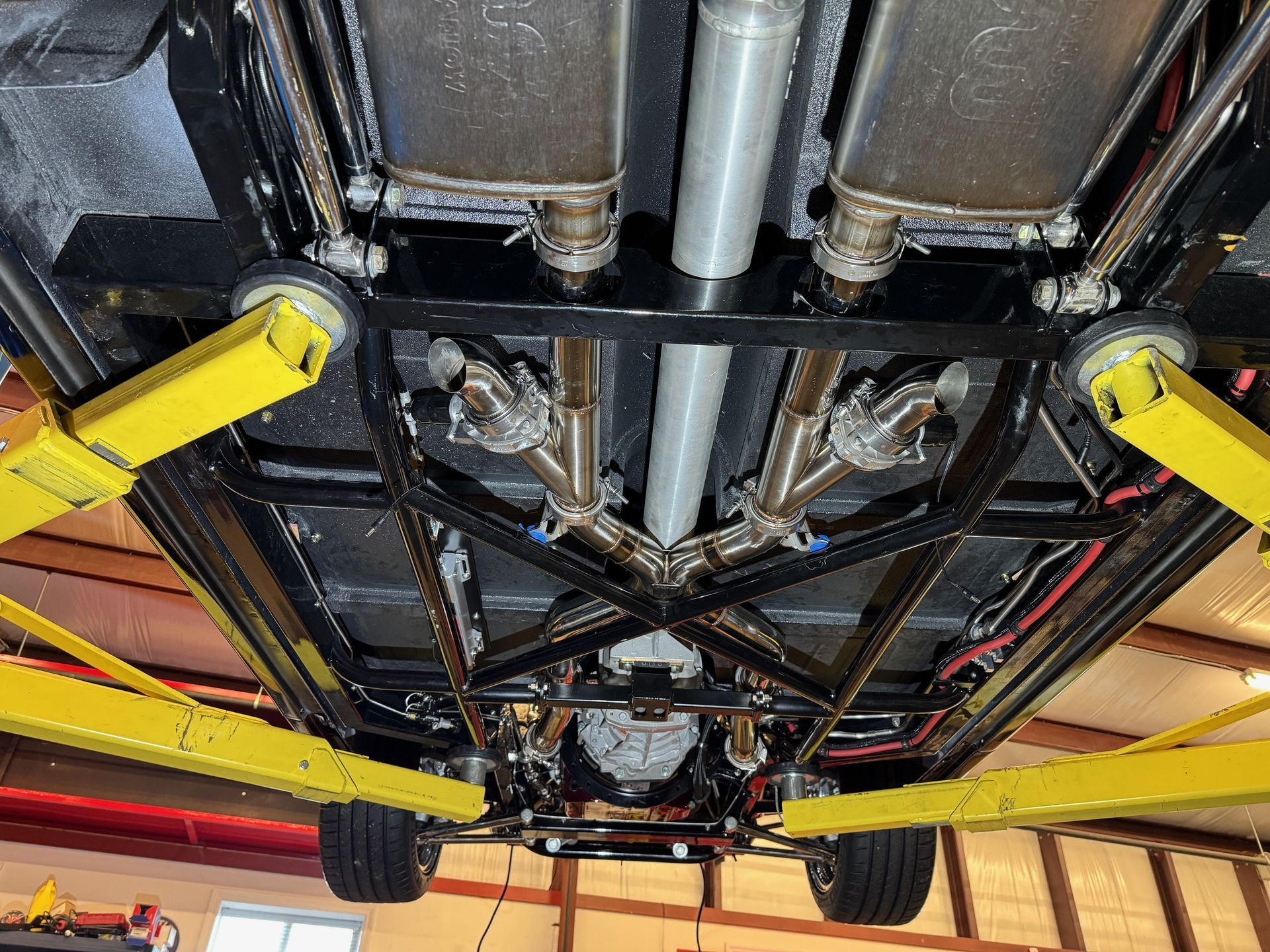 Underbody view of a vehicle lifted on yellow garage stands, showing the black frame, drivetrain, and exhaust system.