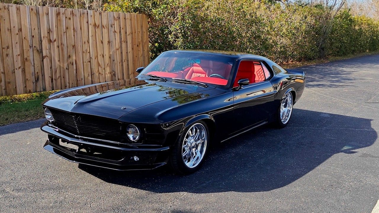 A black 1969 Ford Mustang with a vibrant red interior parked on an asphalt driveway next to a wooden fence.