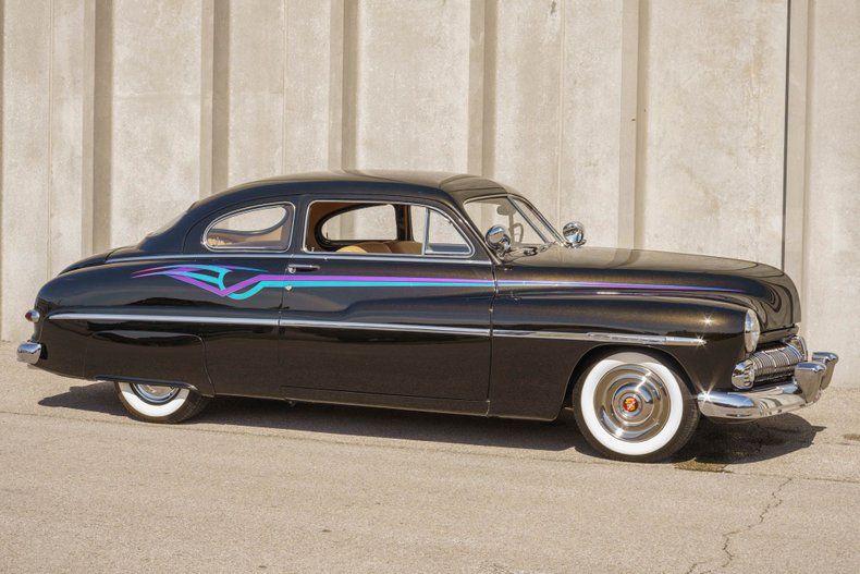A vintage black custom Mercury coupe with a lowered suspension, whitewall tires, and decorative side graphics.