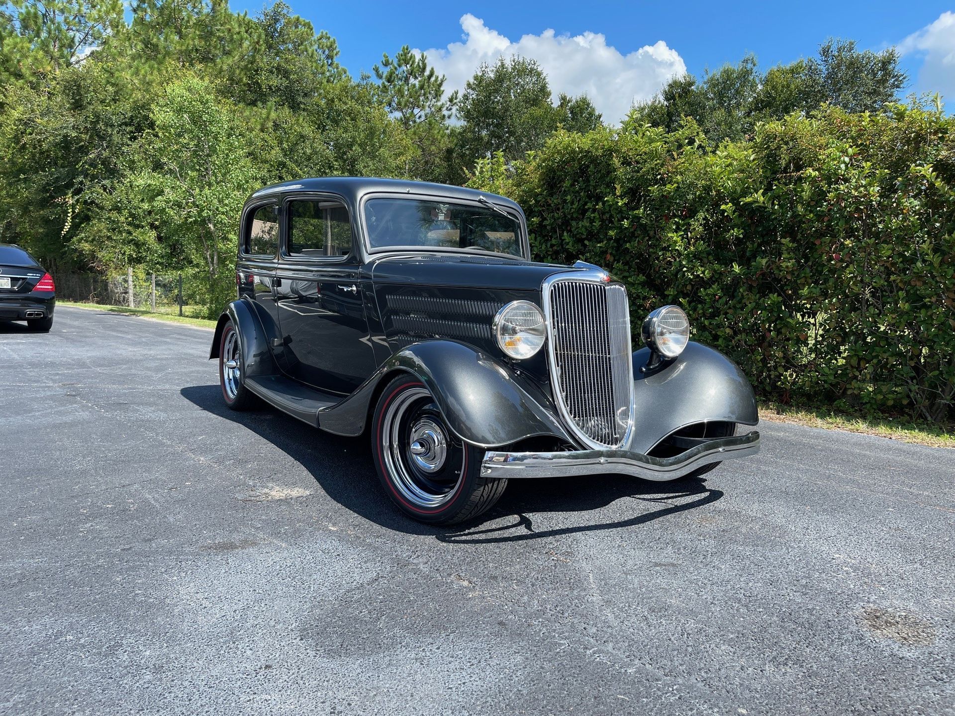 A dark gray vintage Ford coupe parked on a paved driveway surrounded by trees and green bushes.