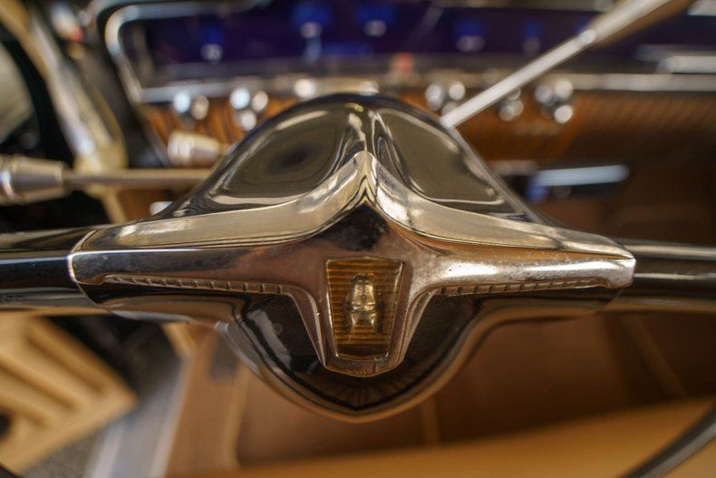 Close-up of a vintage, chrome-accented steering wheel center featuring a small, golden emblem in an interior car setting.